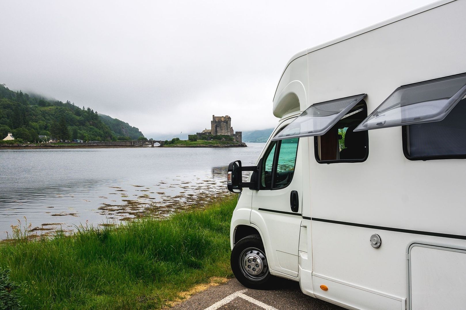 Weißes Wohnmobil am Flussufer mit Burg und bewaldeten Bergen